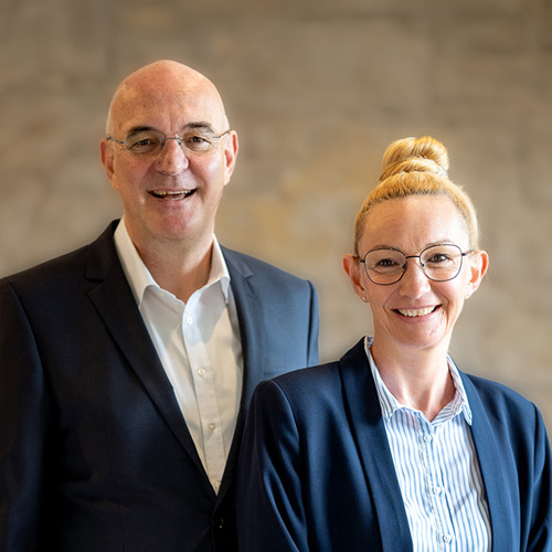 Olaf und Iris Greshake lächeln in dunkler Business-Kleidung in die Kamera. Beide tragen eine Brille. Die Frau hat hochgesteckte, blonde Haare.