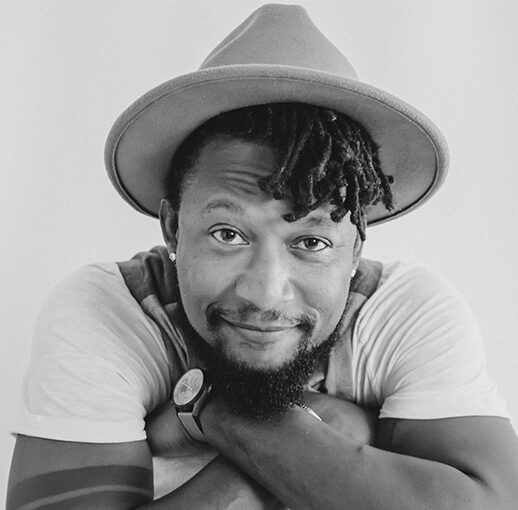 Schwarz-weißes Portraitfoto von Klyive mit Bart, Dreadlocks und Hut. Er lächelt mit verschränkten Armen in die Kamera.